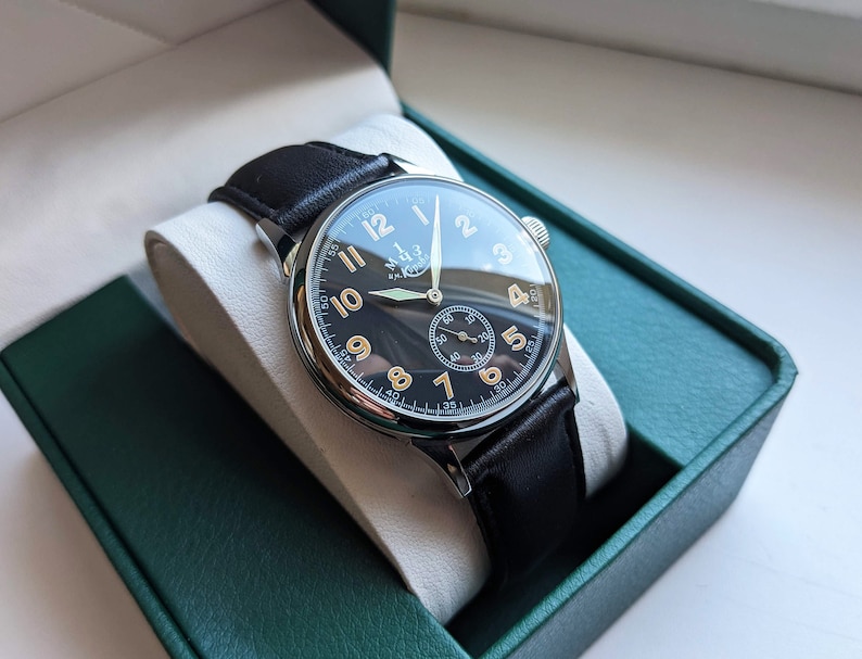 May include: A black-faced wristwatch with gold numerals and hands, a black leather band, and a silver case. The watch is in a green box.
