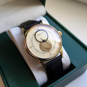 May include: A gold-tone wristwatch with a black leather band. The watch face is white with black markings and features a unique moon phase display. The watch is in a green box.