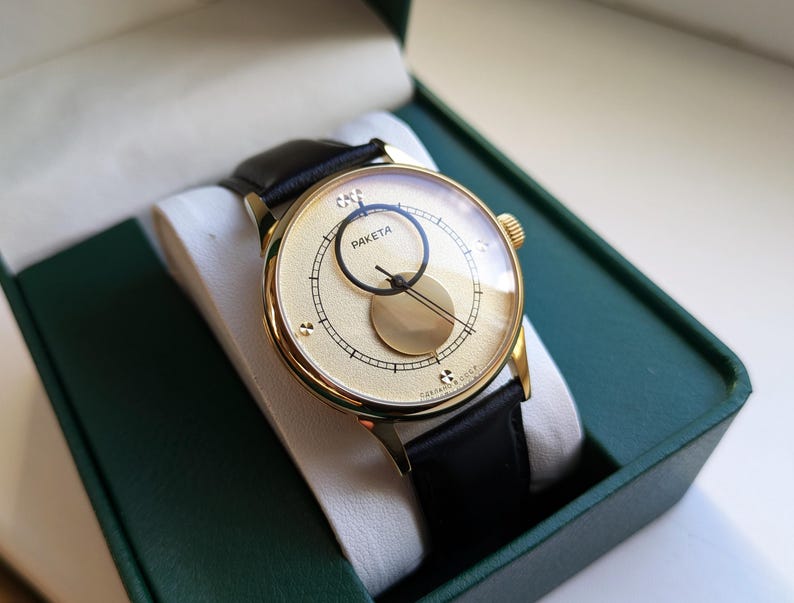 May include: A gold-tone wristwatch with a black leather band. The watch face is white with black markings and features a unique moon phase display. The watch is in a green box.