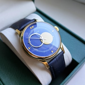 May include: A gold-toned wristwatch with a blue face and a dark blue leather band. The watch face features a circular design with a light-colored circle and a silver-colored ring. The watch is presented in a green box.