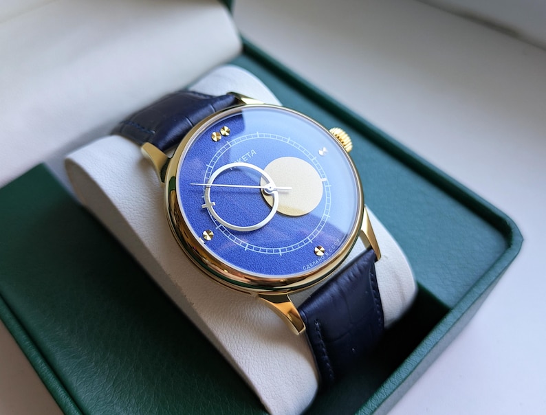 May include: A gold-toned wristwatch with a blue face and a dark blue leather band. The watch face features a circular design with a light-colored circle and a silver-colored ring. The watch is presented in a green box.
