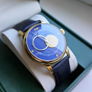 May include: A gold-toned wristwatch with a blue face and a black leather band. The watch face features a circular design with a gold-colored moon shape and a silver hand. The watch is presented in a green box.