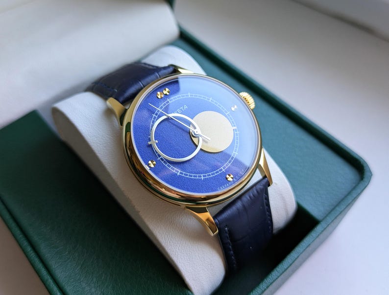 May include: A gold-toned wristwatch with a blue face and a black leather band. The watch face features a circular design with a gold-colored moon shape and a silver hand. The watch is presented in a green box.