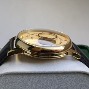 May include: A gold-tone wristwatch with a black leather band. The watch face is white with black markings and a small, round window at the 12 o'clock position.