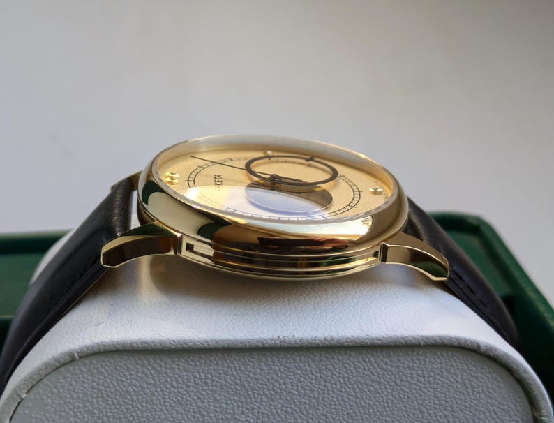 May include: A gold-tone wristwatch with a black leather band. The watch face is white with black markings and a small, round window at the 12 o'clock position.