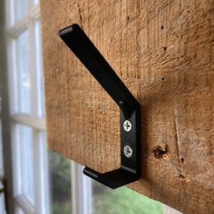 Puede incluir: Un gancho de metal negro con dos tornillos fijados a una superficie de madera. El gancho está inclinado y tiene una punta afilada.