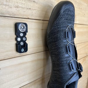 May include: A black cycling shoe with a gray knit upper and a black cleat holder mounted on a wooden surface. The shoe has a dial closure system and multiple straps. The cleat holder has a metal plate with the letters "ENG".
