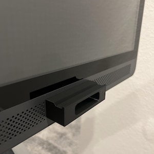 May include: A black, rectangular device attached to the bottom of a black monitor. The device has a slot and a ridged top. The monitor has a perforated speaker grill along the bottom edge.