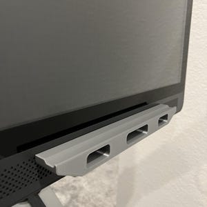 May include: A gray, rectangular bracket with three openings, attached to the bottom of a black screen. The bracket appears to be made of plastic or a similar material. The screen is mounted on a stand.