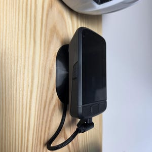 May include: A black cycling computer mounted on a wooden surface with a black cord. A white bicycle helmet is visible above the computer. The computer is attached to a black mount.