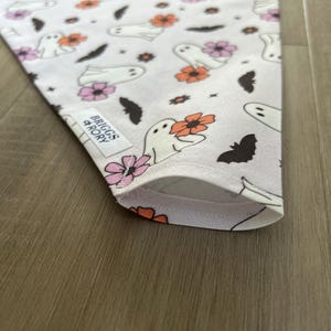 K&ouml;nnte beinhalten: Ein wei&szlig;es Bandana mit einem sich wiederholenden Muster aus Geistern, Flederm&auml;usen und Blumen in Rosa-, Orange- und Braunt&ouml;nen. Das Bandana hat ein wei&szlig;es Etikett mit dem Text "Briggs & Rory" in Schwarz.