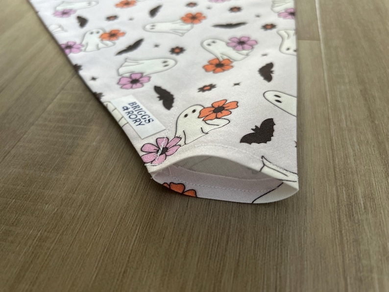K&ouml;nnte beinhalten: Ein wei&szlig;es Bandana mit einem sich wiederholenden Muster aus Geistern, Flederm&auml;usen und Blumen in Rosa-, Orange- und Braunt&ouml;nen. Das Bandana hat ein wei&szlig;es Etikett mit dem Text "Briggs & Rory" in Schwarz.