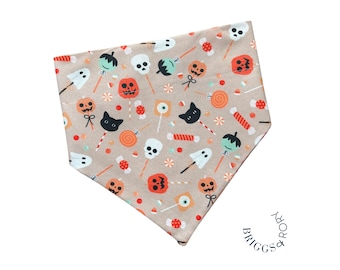 May include: A tan bandana with a Halloween-themed print featuring ghosts, pumpkins, candy, and skulls. The bandana has a scalloped edge and is decorated with a black and white "Briggs & Rook" logo.