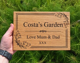Puede incluir: Un letrero rectangular de madera clara con las palabras "Costa's Garden" y "Love Mum & Dad" grabadas en negro. Cuenta con adornos florales en las esquinas y un borde.