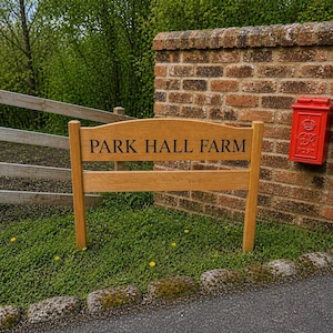 May include: A wooden sign reading "PARK HALL FARM" is positioned in front of a brick wall with a red postbox. The sign is made of light brown wood and is set in a grassy area.