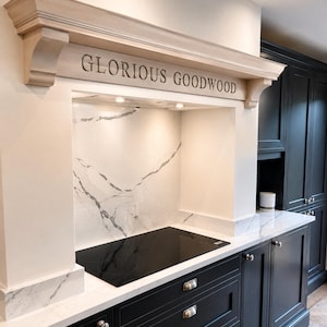 May include: A kitchen range hood featuring a light wood mantel with the text GLORIOUS GOODWOOD carved into the front. The hood is positioned above a black induction cooktop and a white marble backsplash with grey veining, set against black cabinetry.