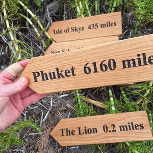 May include: Wooden directional signs with black text indicating distances to various locations. One sign reads "Phuket 9913 km". Other signs point to "Isle of Skye 700 km" and "The Lion 0.3 km". The signs are held over green foliage.