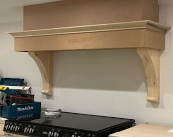 May include: A light brown wooden kitchen range hood with a black glass cooktop and a cream-colored oven with black accents. The hood has a decorative curved design and is mounted above the stove.
