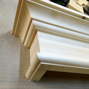 May include: A close-up of a wooden shelf bracket with a decorative edge. The bracket is made of unfinished pine wood and has a simple, elegant design.