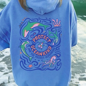 May include: A light blue hoodie with a graphic on the back that says "Protect Oceankind and You Protect Humankind." The graphic features a dolphin, shark, turtle, and octopus in shades of green, pink, and blue. The hoodie is worn at the beach.