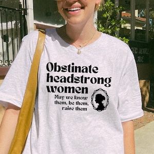 May include: A grey t-shirt with black text that reads "Obstinate headstrong women. May we know them, be them, raise them."  The text is in a retro font style. A black silhouette of a woman's head is in a circle above the text.