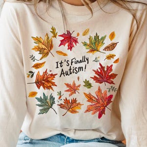 Può includere: Maglietta a maniche lunghe color crema con un design di foglie colorate e la scritta "It's Finally Autism!". Le foglie sono nei toni del rosso, arancione, giallo e verde. La maglietta è indossata da una persona.