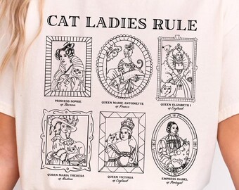 Op de afbeelding: Een wit T-shirt met de tekst "CAT LADIES RULE" gedrukt op de voorkant. Het shirt heeft zwart-witte illustraties van historische figuren met katten, waaronder Prinses Sophie van Beieren, Koningin Marie Antoinette, Koningin Elizabeth I, Koningin Maria Theresia van Oostenrijk, Koningin Victoria en Keizerin Isabel van Brazilië.