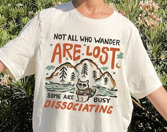 May include: White t-shirt with a graphic of a raccoon in a forest with the text "Not all who wander are lost. Some are busy dissociating."