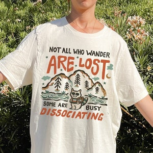 May include: White t-shirt with a graphic of a raccoon in a forest with the text "Not all who wander are lost. Some are busy dissociating."