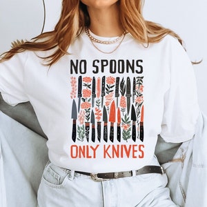May include: White long-sleeved t-shirt with a graphic of black knives and red flowers. The text "NO SPOONS ONLY KNIVES" is printed on the shirt.