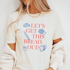 Può includere: Una maglietta bianca con una grafica blu e rossa che dice "Let's Get This Bread, Oui?" con illustrazioni di pane.