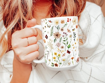 Può includere: Una tazza in ceramica bianca con un motivo floreale che presenta una varietà di fiori e farfalle. La tazza è tenuta da una mano.