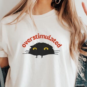 Puede incluir: Camiseta color crema con la palabra "overstimulated" en rojo sobre un gráfico de gato negro con ojos amarillos. El gato asoma por debajo del texto. La camiseta se lleva con una chaqueta vaquera.