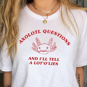 Peut inclure: T-shirt blanc avec un graphique d'axolotl rouge et le texte "AXOLOTL QUESTIONS AND I'LL TELL A LOT'O'LIES".