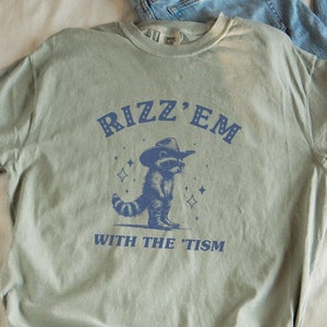Peut inclure: Un t-shirt vert avec un raton laveur bleu portant un chapeau de cowboy et le texte "Rizz 'Em With The 'Tism" imprim&eacute; dessus.