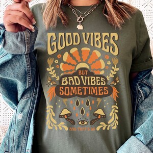 May include: Green t-shirt with a retro graphic design that says "Good Vibes But Bad Vibes Sometimes And That's Ok". The graphic features a sun, flowers, mushrooms, and a third eye.
