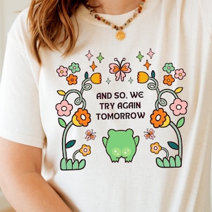 May include: A white t-shirt with a colourful graphic design featuring flowers, a butterfly, and a frog. The text on the shirt reads "And so, we try again tomorrow."