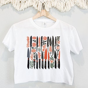 May include: White cropped t-shirt with a graphic print of black knives and orange flowers.