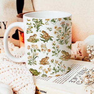 May include: White ceramic mug with a floral pattern featuring green leaves, white flowers, and brown frogs. The mug has a white handle.