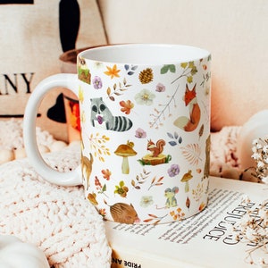 May include: White ceramic mug with a autumn-themed design featuring woodland animals, leaves, and mushrooms. The mug has a large handle and is sitting on a stack of books.