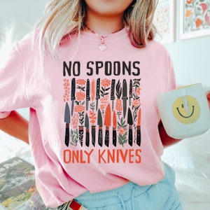 May include: Pink t-shirt with a graphic of knives and the text "No Spoons Only Knives".