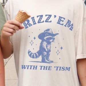 Peut inclure: Deux personnes portant des t-shirts avec un raton laveur portant un chapeau de cowboy et le texte "Rizz'em with the 'tism".