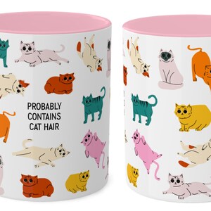 May include: White ceramic mug with a pink rim. The mug features a repeating pattern of colourful cartoon cats in various poses. The text "PROBABLY CONTAINS CAT HAIR" is printed in black on the mug.