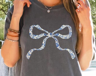 May include: A gray t-shirt with a white bow printed on the front. The bow is decorated with a blue and white floral pattern.