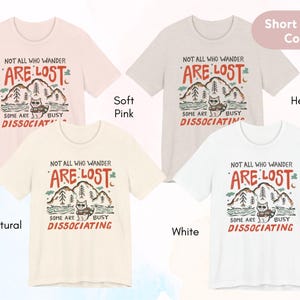 May include: Four short-sleeved t-shirts with a graphic print of a cat in a forest with the text "Not all who wander are lost. Some are busy dissociating."