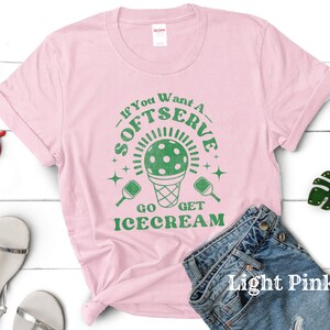 以下が含まれることがあります： 薄いピンクのTシャツに、緑色のピクルボールのパドルとアイスクリームコーンのグラフィックが描かれています。Tシャツのテキストは「If you want a soft serve go get ice cream」です。