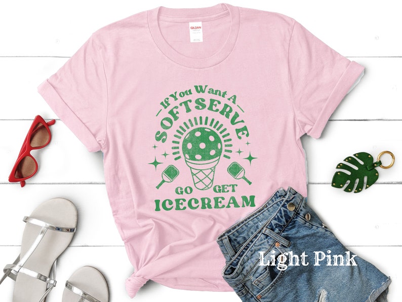 以下が含まれることがあります： 薄いピンクのTシャツに、緑色のピクルボールのパドルとアイスクリームコーンのグラフィックが描かれています。Tシャツのテキストは「If you want a soft serve go get ice cream」です。