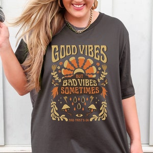 May include: A dark gray t-shirt with a retro-style graphic design. The graphic features a sun with orange rays, flowers, butterflies, and the text "Good Vibes But Bad Vibes Sometimes And That's Ok."