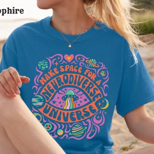 May include: A blue t-shirt with a psychedelic graphic design featuring a rainbow eye, stars, planets, and the text "Make space for a neurodiverse universe".
