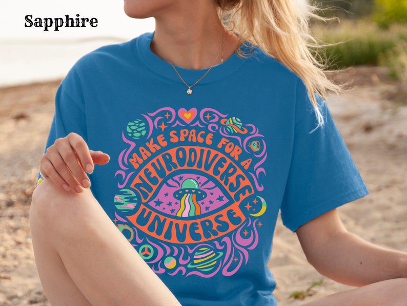 May include: A blue t-shirt with a psychedelic graphic design featuring a rainbow eye, stars, planets, and the text "Make space for a neurodiverse universe".